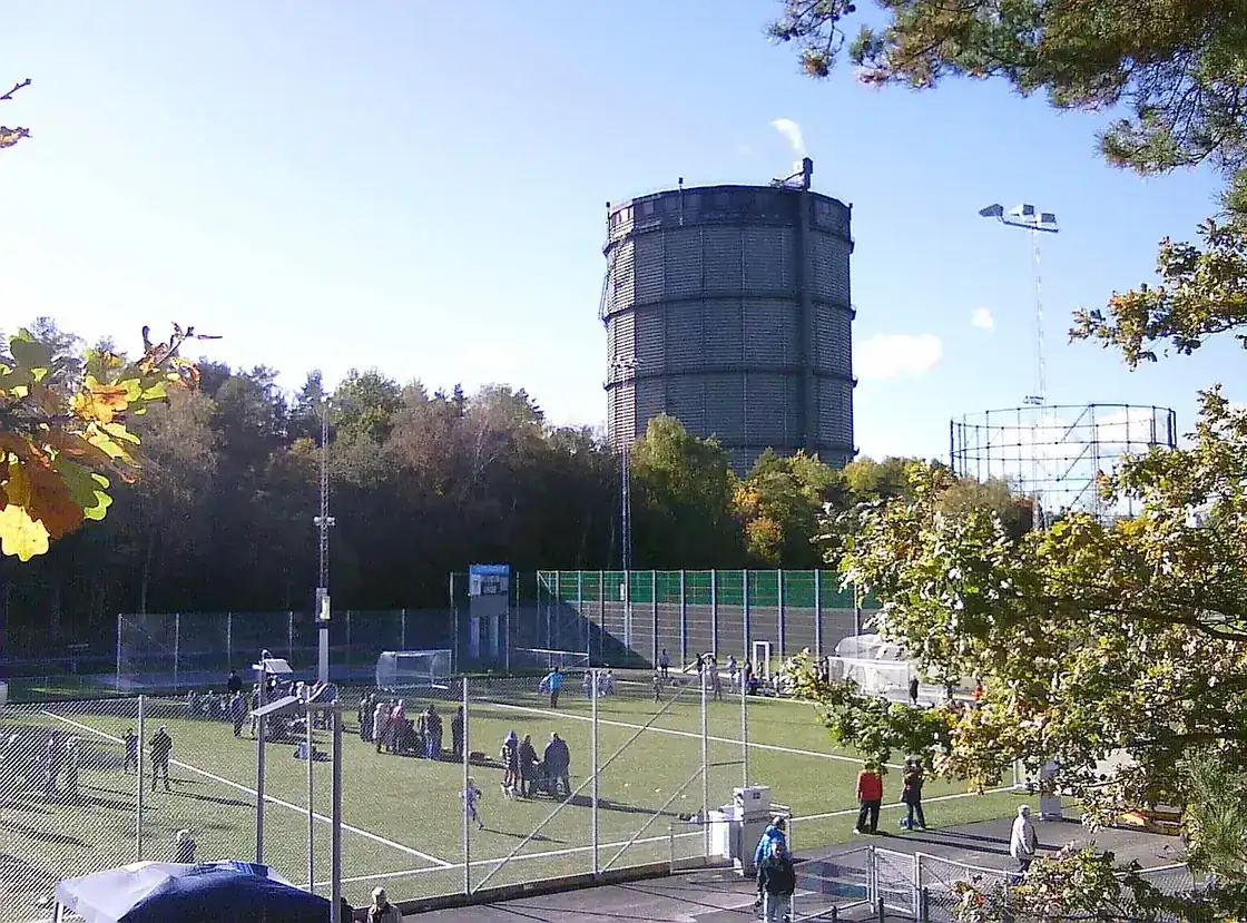 Konstgräsplan omgiven av träd med gasklockan i Hjorthagen i bakgrunden